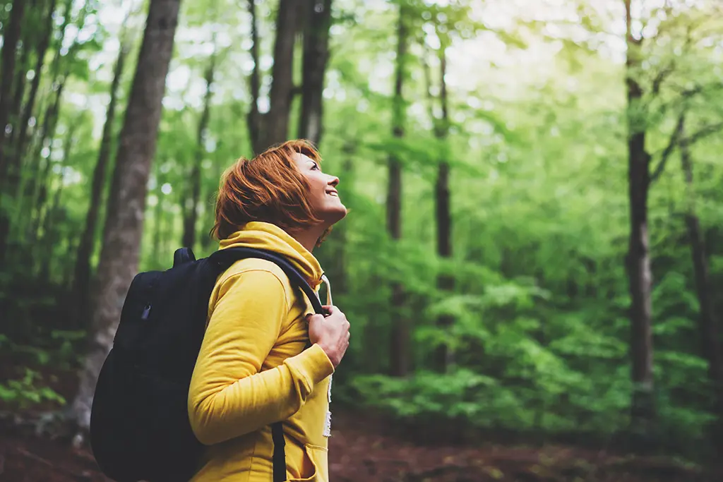 Woman with red hair wearing yellow jacket explores peaceful green forest, symbolizing Evergreen Energy Partners' new brand refresh and identity, and their expanded offerings that make energy efficiency initiatives accessible to all.